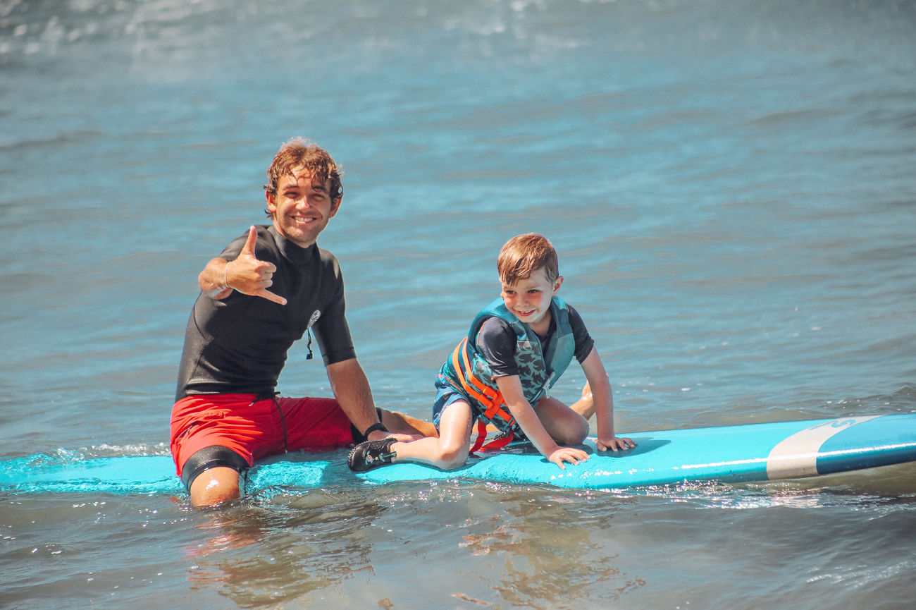 A Local Pros Maui surf instructor sits on a surfboard with a smiling child, learning how to surf near Kaanapali.