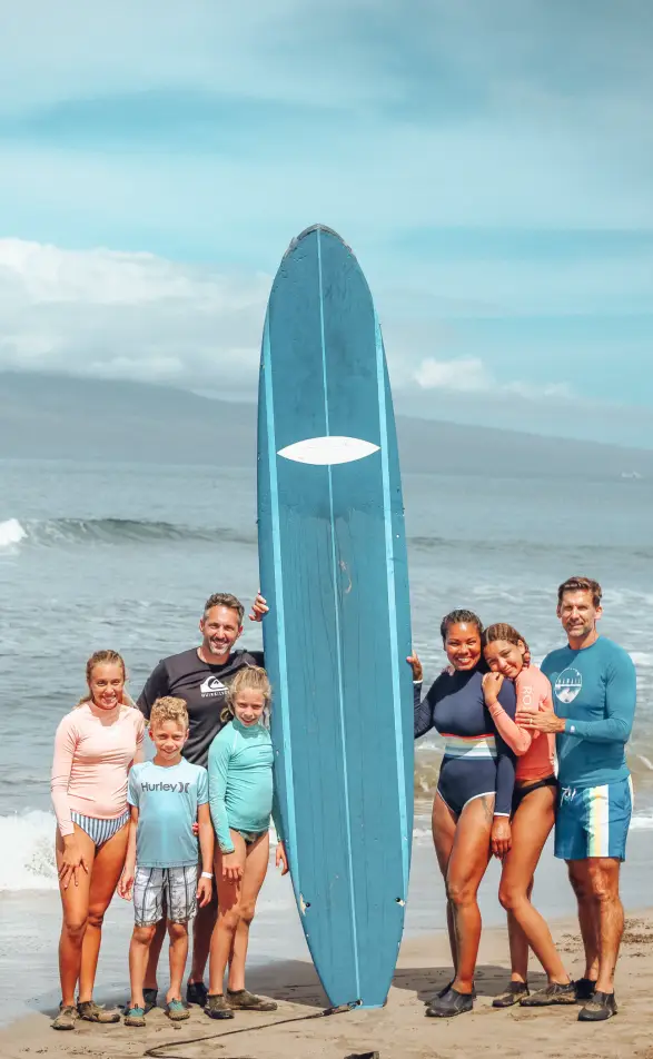 A family of six posing on the beach with a tall blue surfboard after a surf lesson in Maui