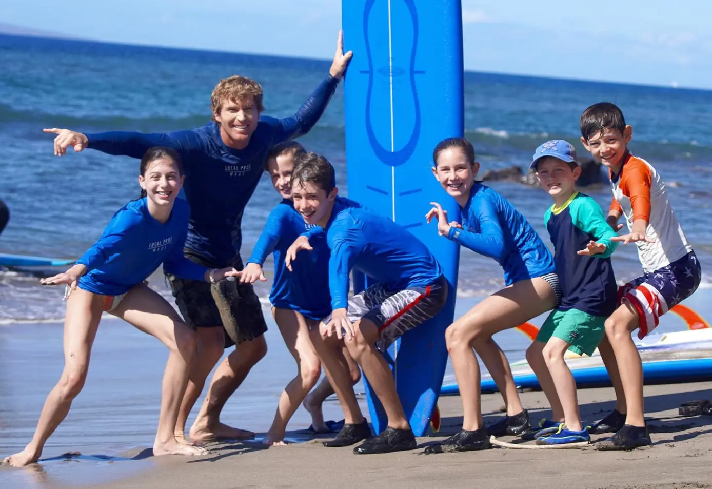 A surf instructor and a group of children taking a group surfing lesson pose for the camera on shore. 