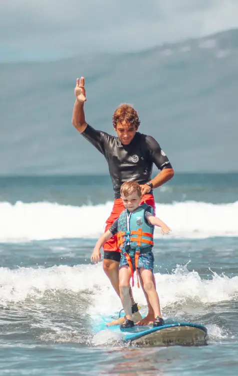 A surf instructor supports a young surfing student in Lahaina, helping him balance while catching a wave.