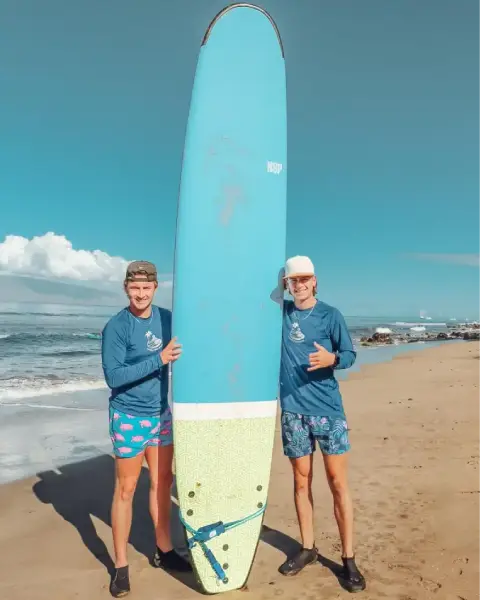 Two Local Pros Maui surf instructors standing on the beach with a blue longboard, smiling and giving shaka signs under a blue sky