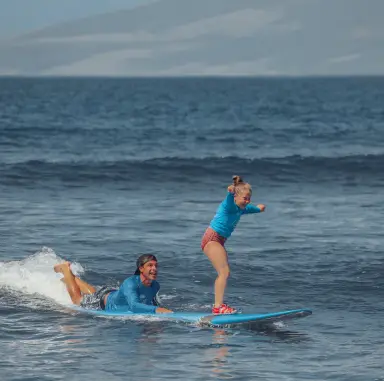 A surf instructor lying on a surfboard in shallow water while a young child stands and surfs a small wave in Maui